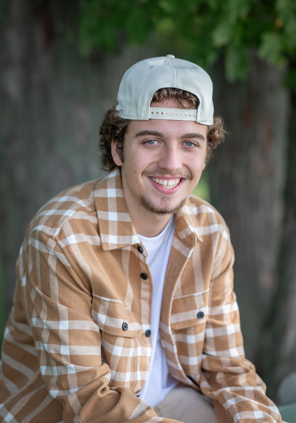 Young man in a tan plaid flannel shirt and backwards white cap smiling outdoors near green trees.