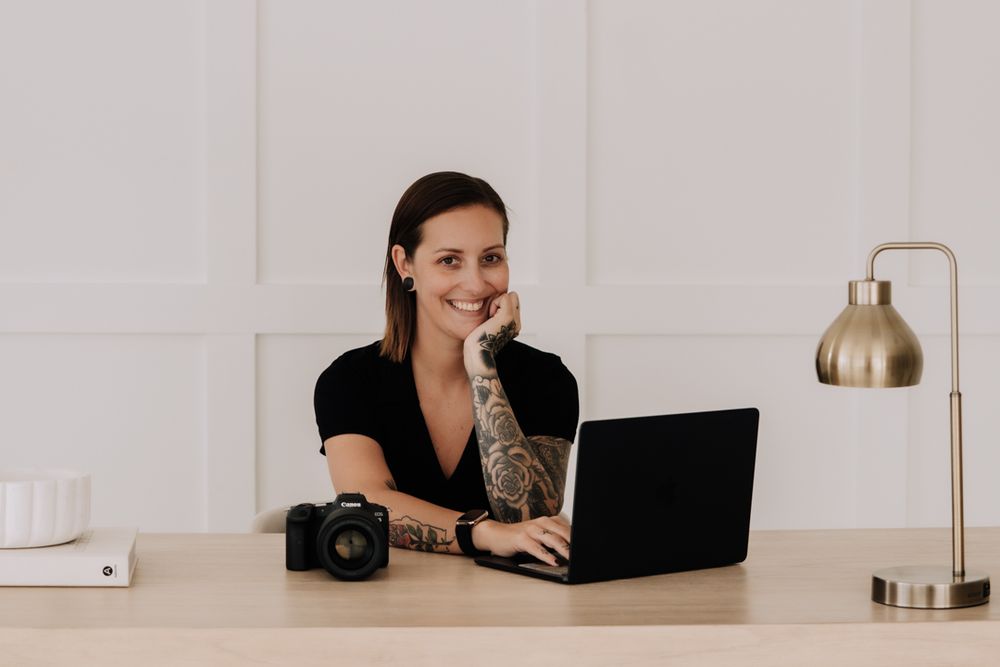 Smiling woman with tattoos sitting at a desk with a laptop and camera in a bright modern home office.