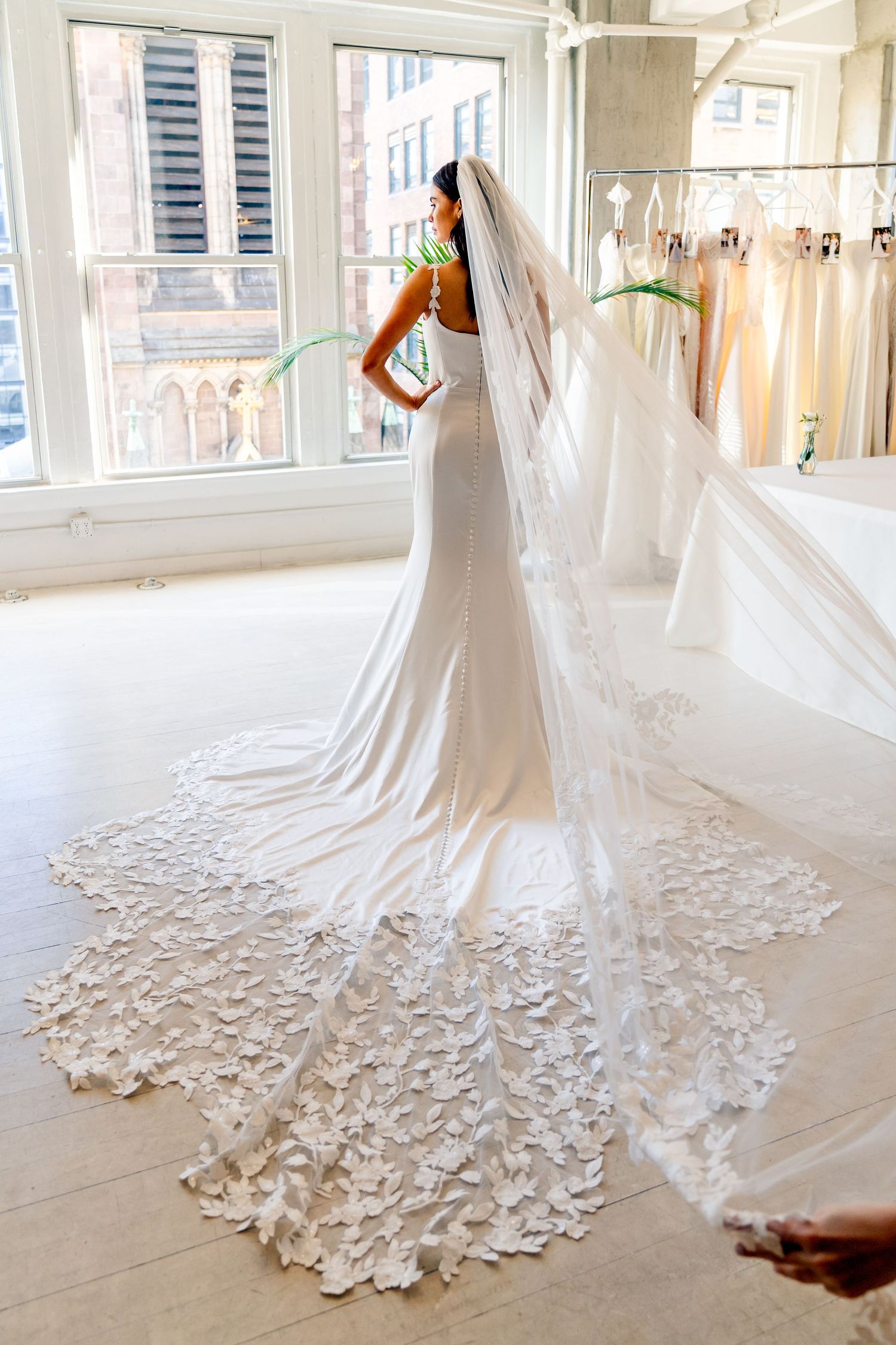 Bride in fitted white gown with floral lace cathedral veil spread on floor in bridal boutique.