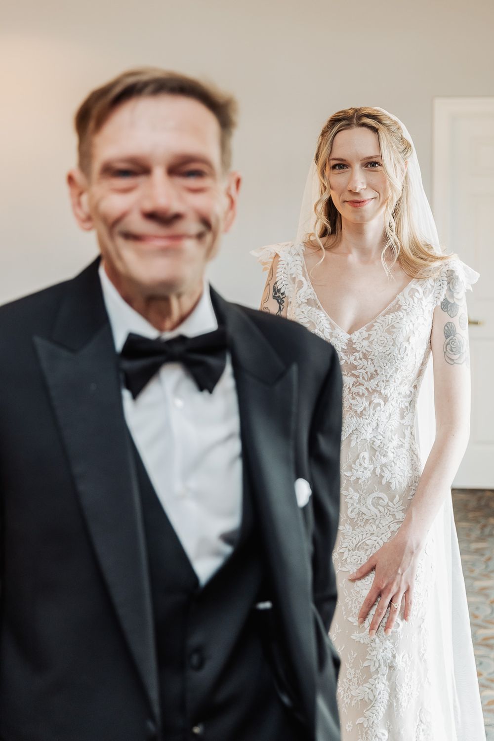A smiling groom in a tuxedo stands in the foreground as a bride in a lace wedding gown poses behind him.