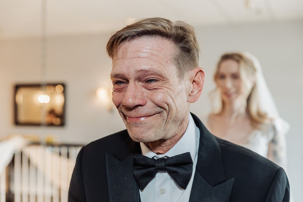 Emotional man in tuxedo with bow tie crying tears of joy at wedding ceremony with bride in background.