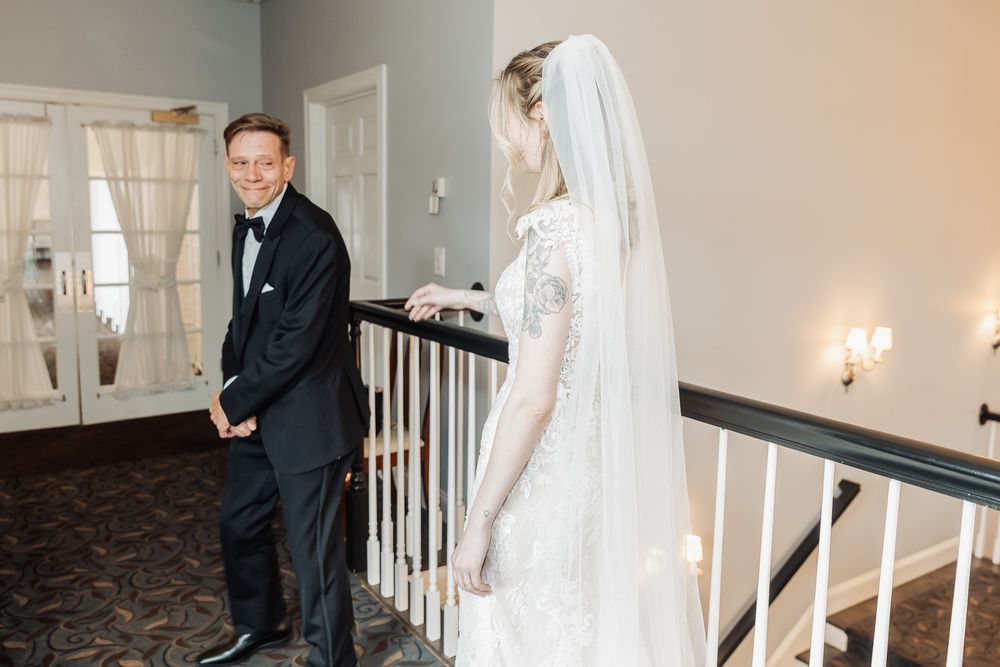Groom in tuxedo smiles at bride in lace wedding dress and veil on elegant staircase during first look.