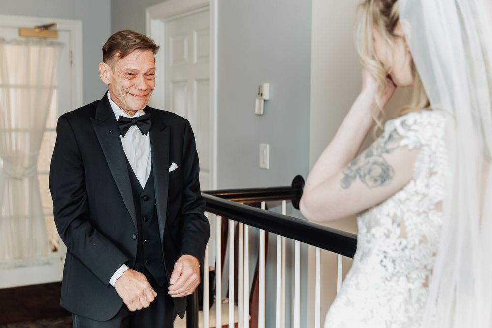 Groom in black tuxedo smiles at bride in lace wedding dress and veil during emotional first look on staircase.