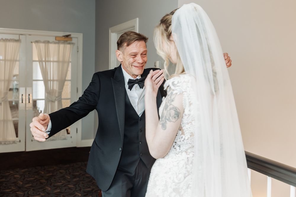 Groom in black tuxedo shares first dance moment with bride in lace wedding dress and veil indoors.