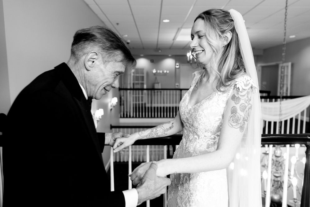 Black and white photo of a bride in lace gown and veil sharing a tender moment with an older gentleman in a tuxedo.