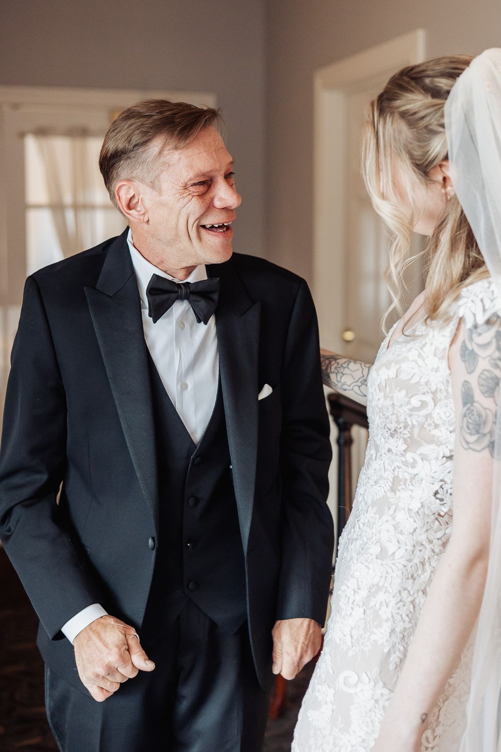 Older man in black tuxedo smiling at bride in lace wedding dress during emotional father-daughter first look moment.