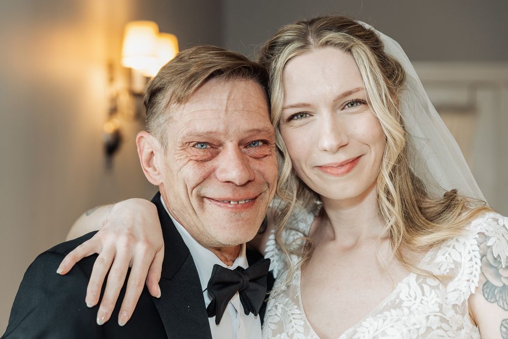A bride in a white lace veil smiles with an older man in a tuxedo at a wedding celebration.