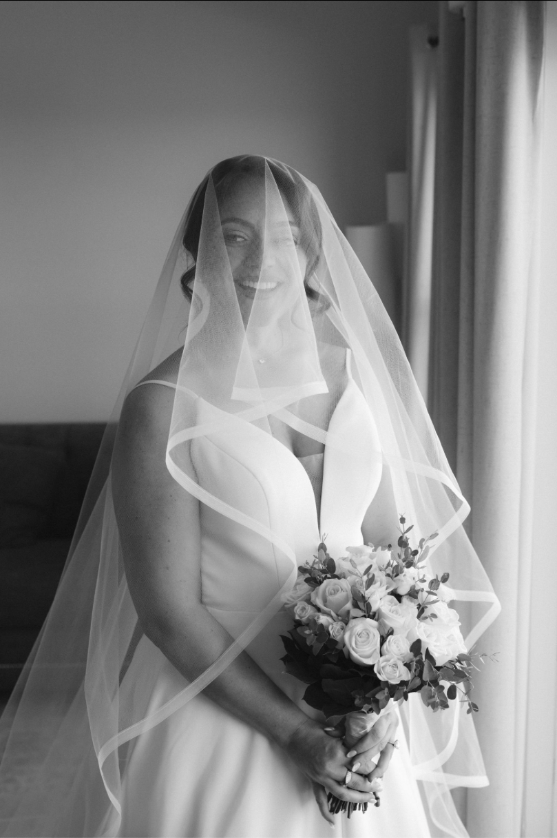 Black and white portrait of a smiling bride in a white gown and veil holding a bouquet of flowers near a window.