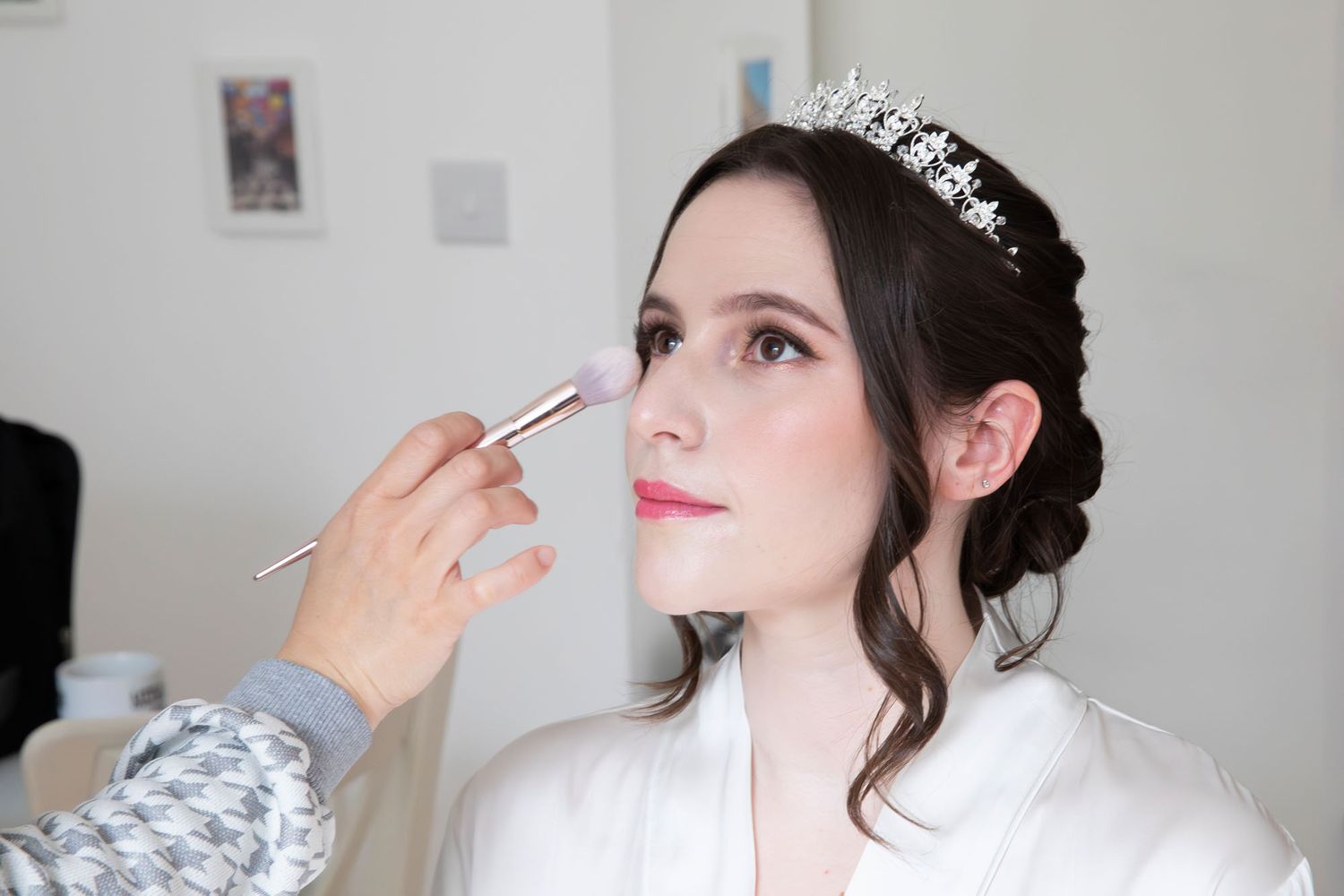 Bride getting makeup applied with a blush brush, wearing a tiara and white robe during bridal preparations.