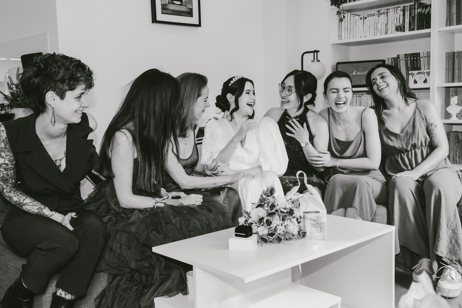 Black and white photo of a bride laughing with bridesmaids on a couch, bouquet on coffee table.