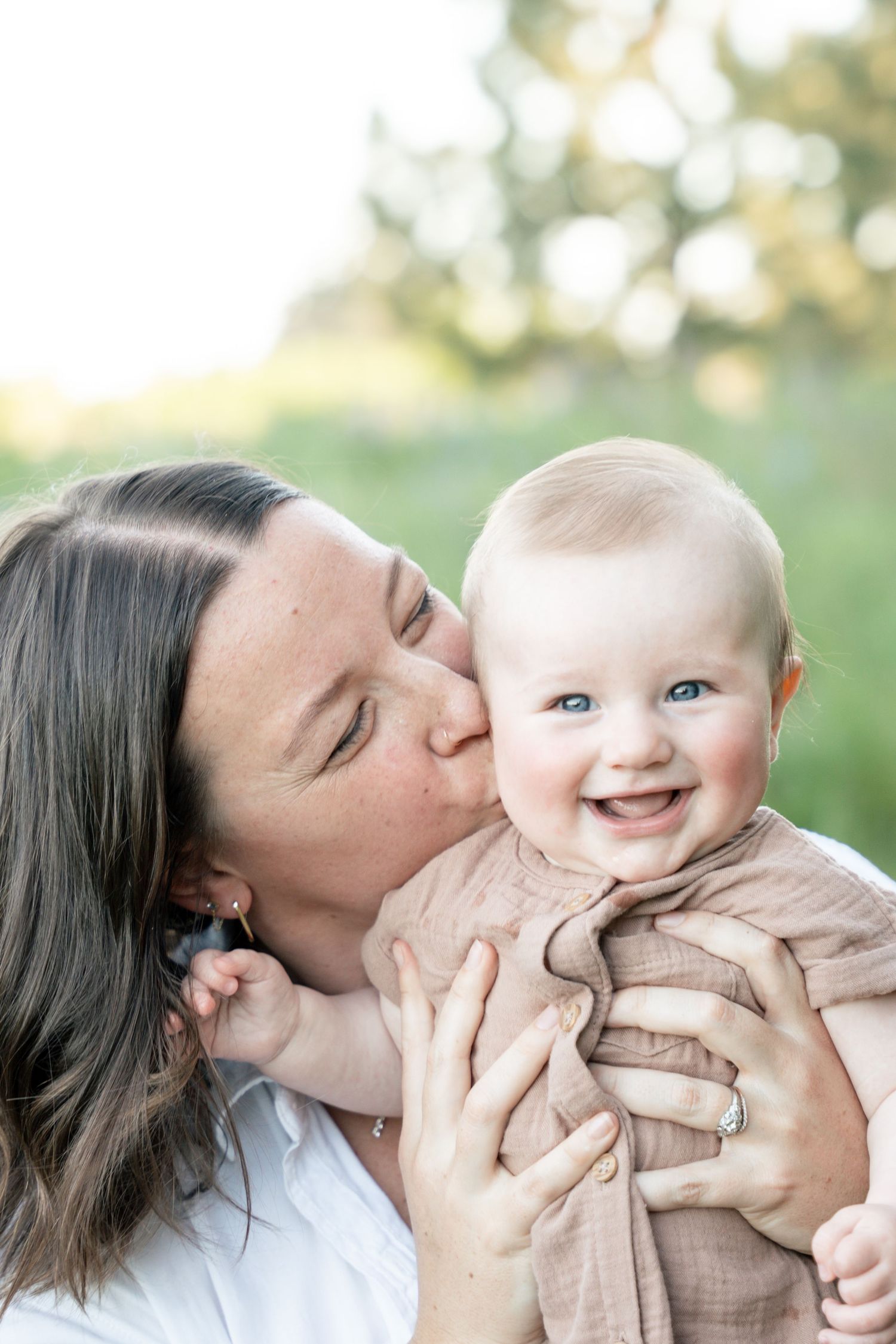 A mother kisses her smiling blue-eyed baby outdoors in a sunny green field.