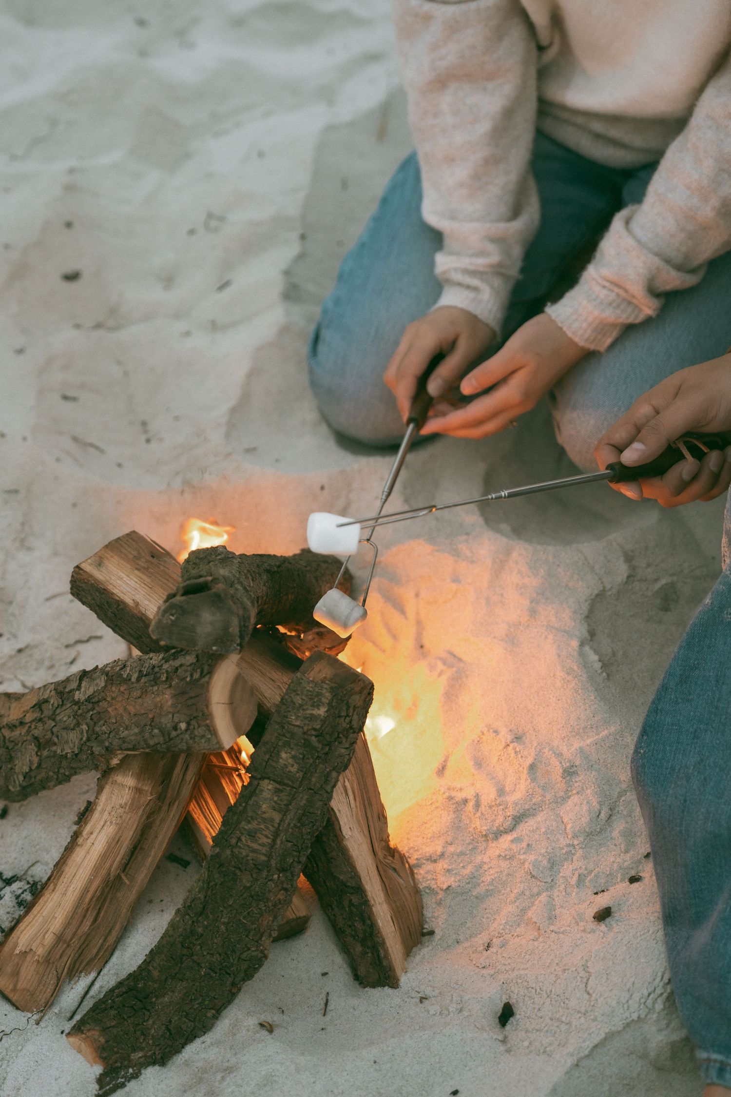 Person roasting marshmallows on sticks over a small campfire on a sandy beach at dusk.