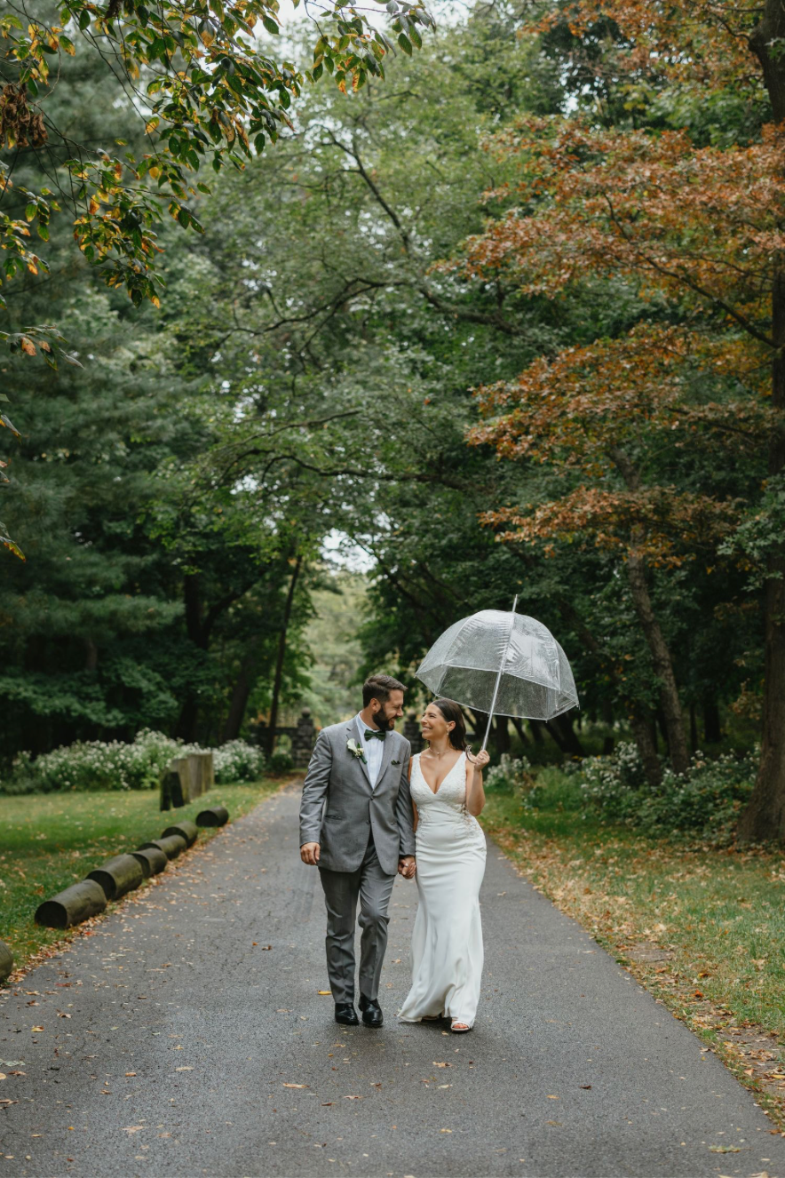 Bride and groom walk under clear umbrella on rainy tree-lined path, bride in white gown, groom in gray suit.