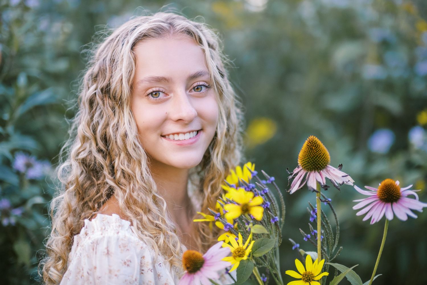 Young woman with curly blonde hair smiling among colorful wildflowers including yellow and pink coneflowers in a garden.