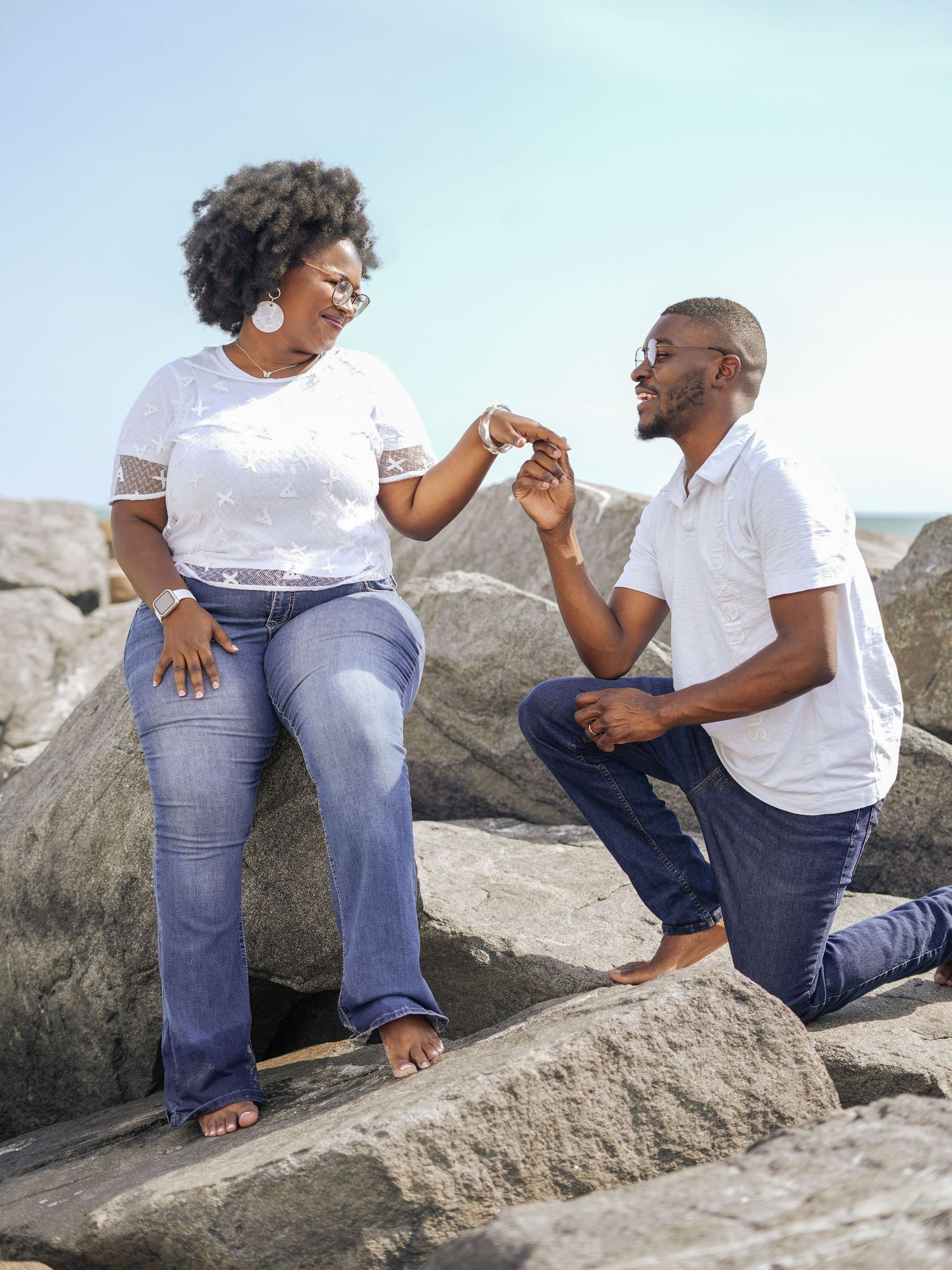 Couple holding hands walking through shallow ocean water near rocks on a beach, wearing white shirts and jeans.