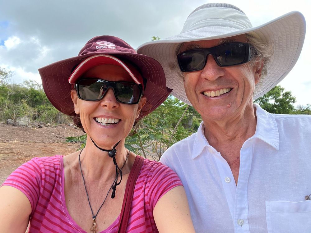 A smiling couple wearing sun hats and sunglasses takes a selfie outdoors in a sunny, tropical landscape.