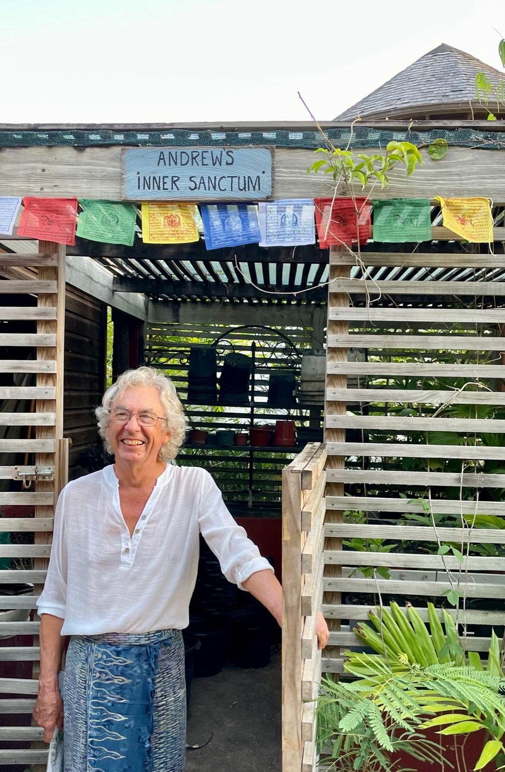 Smiling elderly person standing at entrance of wooden structure with colorful prayer flags and Andrews Inner Sanctum sign.