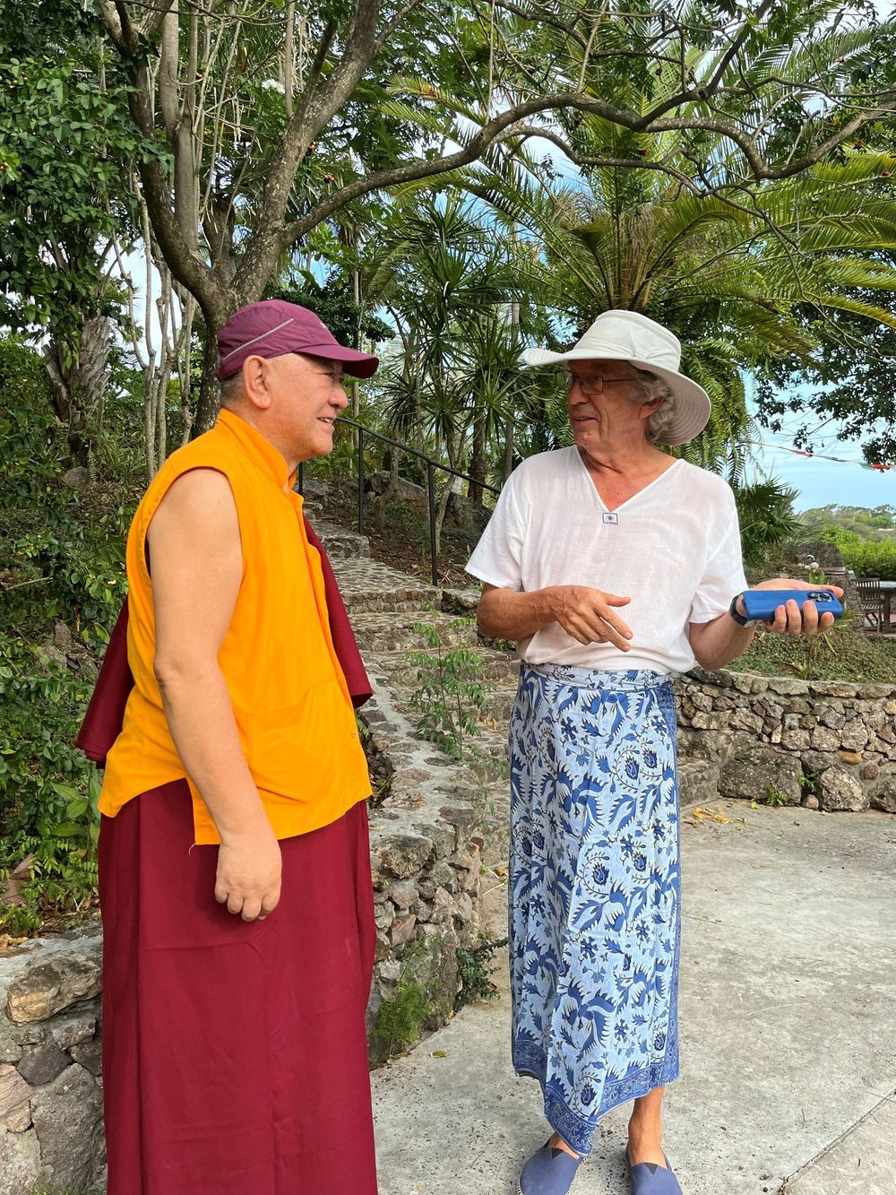 Buddhist monk in yellow robes talks with a woman in a white shirt and floral skirt outdoors near stone wall.