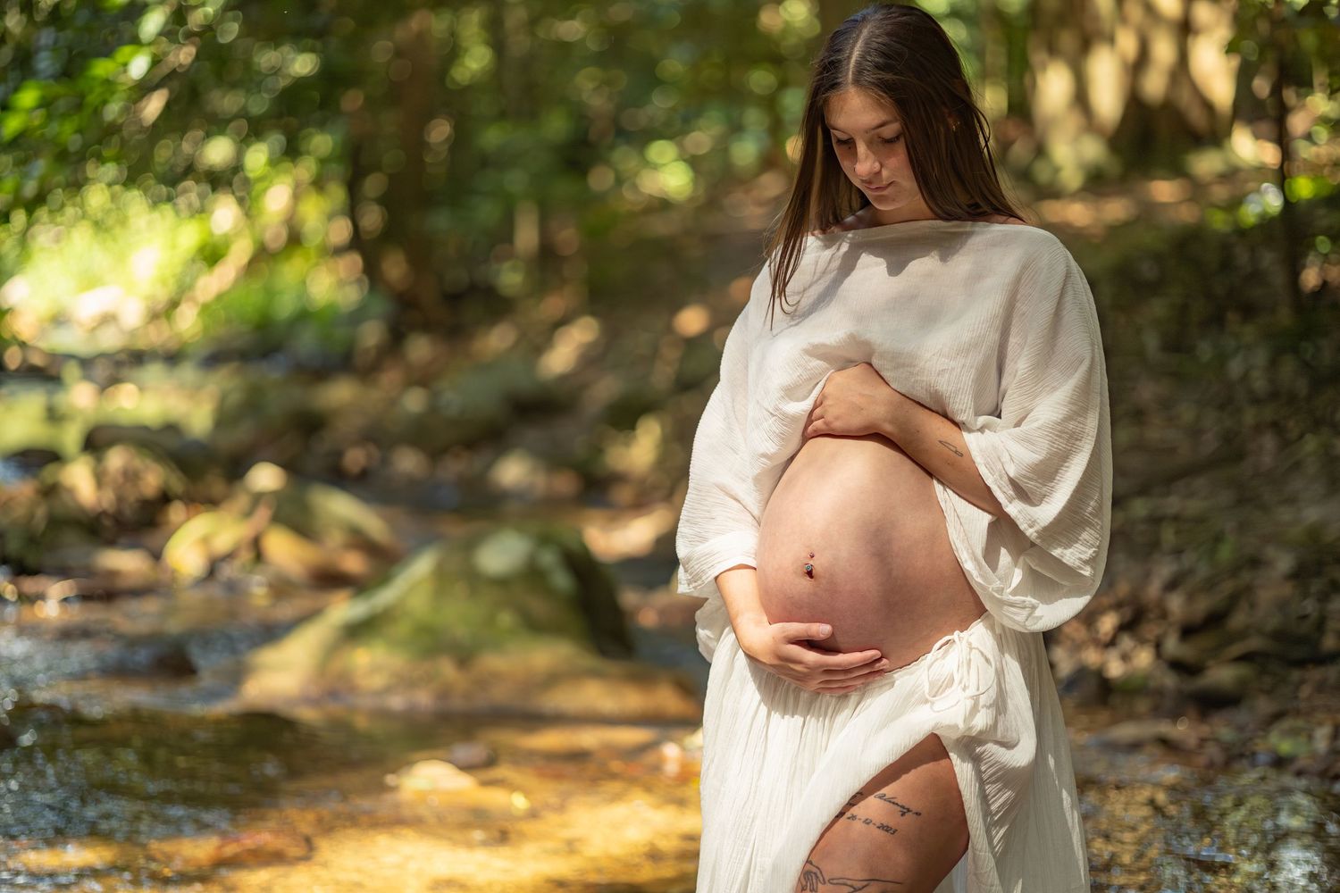 A pregnant woman in a flowing white dress cradles her baby bump while standing near a sunlit forest stream.
