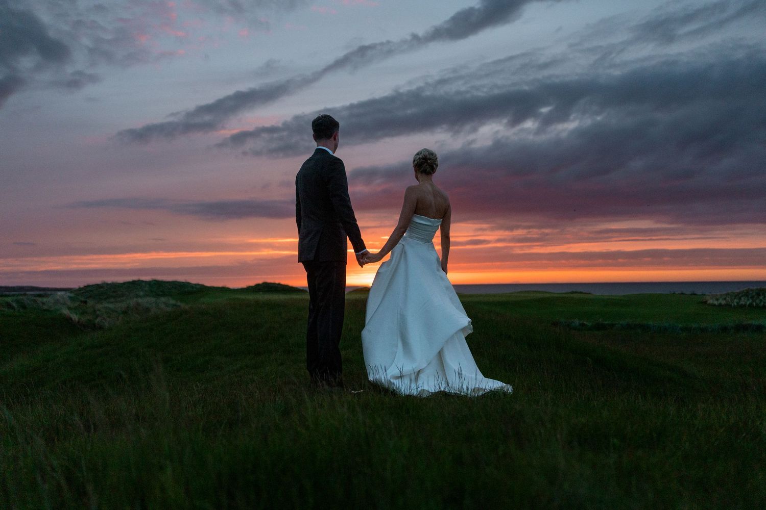 Bride and groom holding hands facing a vibrant sunset over green hills on their wedding day.