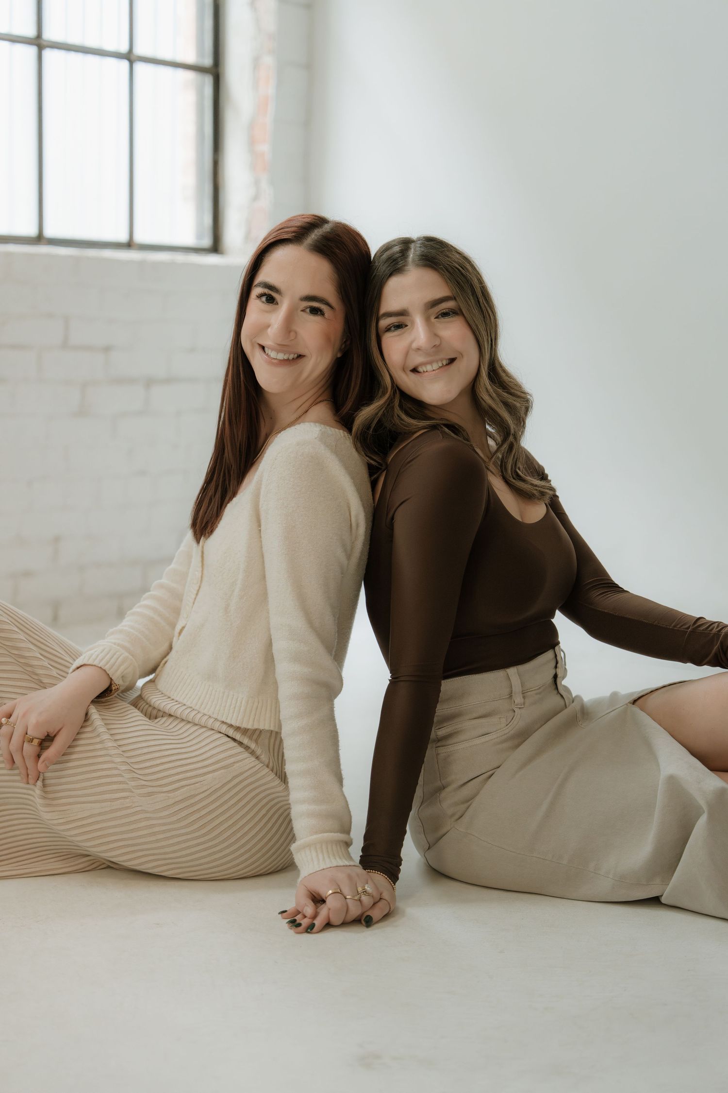 Two smiling young women sitting back-to-back on a white floor, wearing neutral tones, in a bright studio setting.