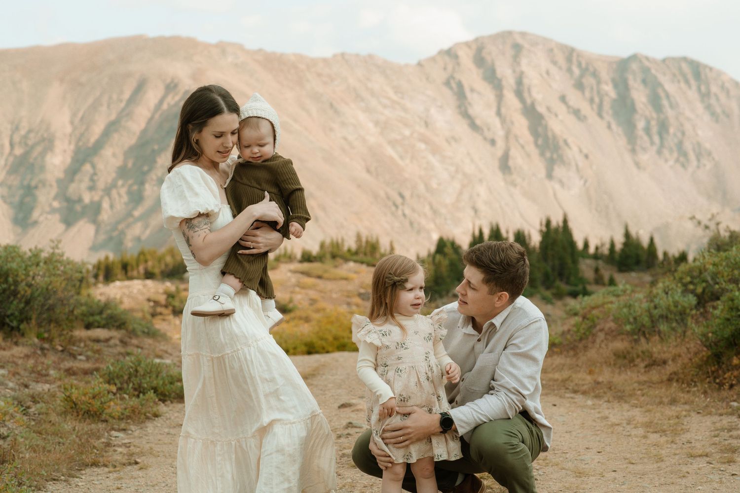 Family of four posing in a mountain meadow with alpine lake and rocky peaks in the background.