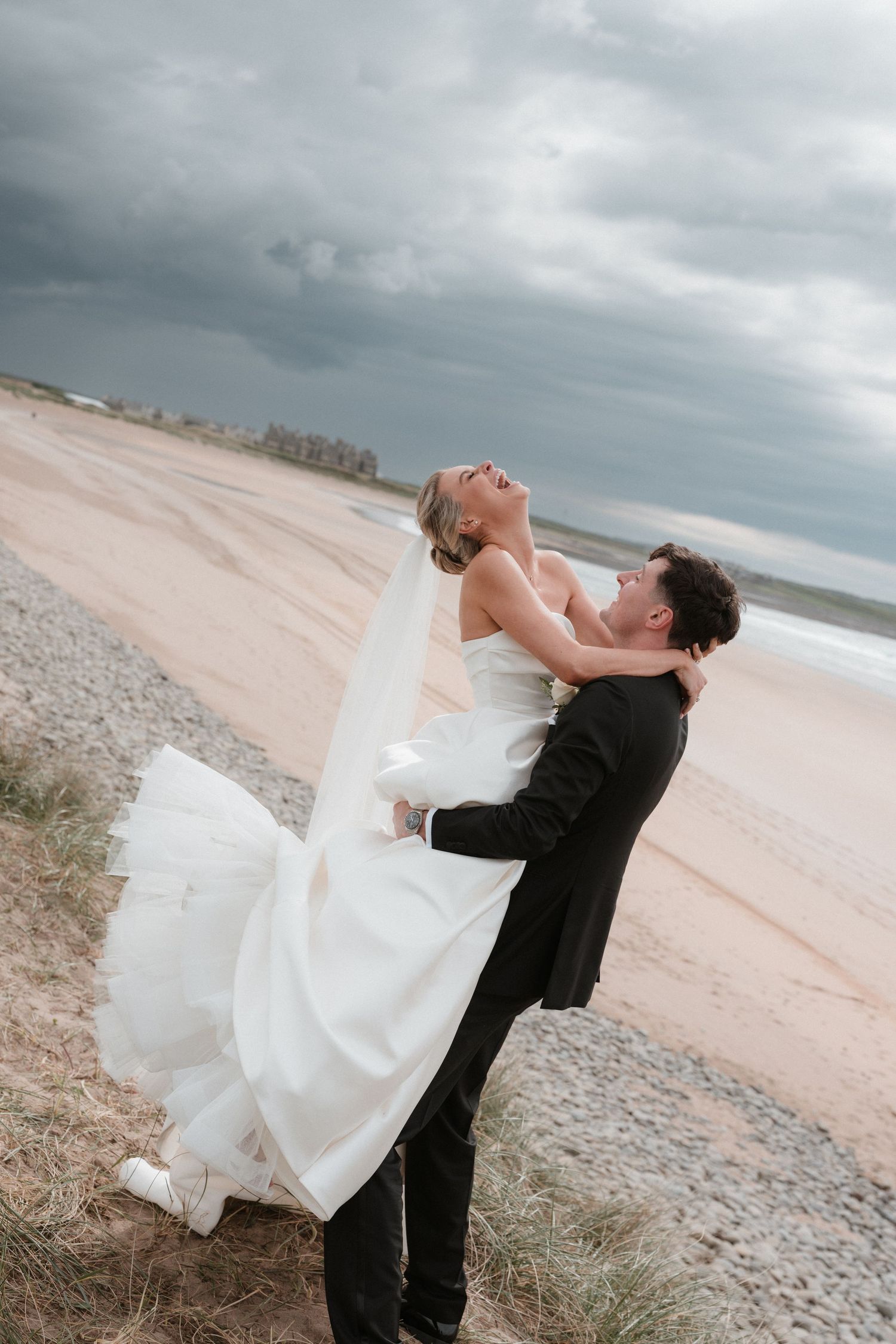 Groom lifting bride in white gown on a sandy beach under dramatic stormy skies.