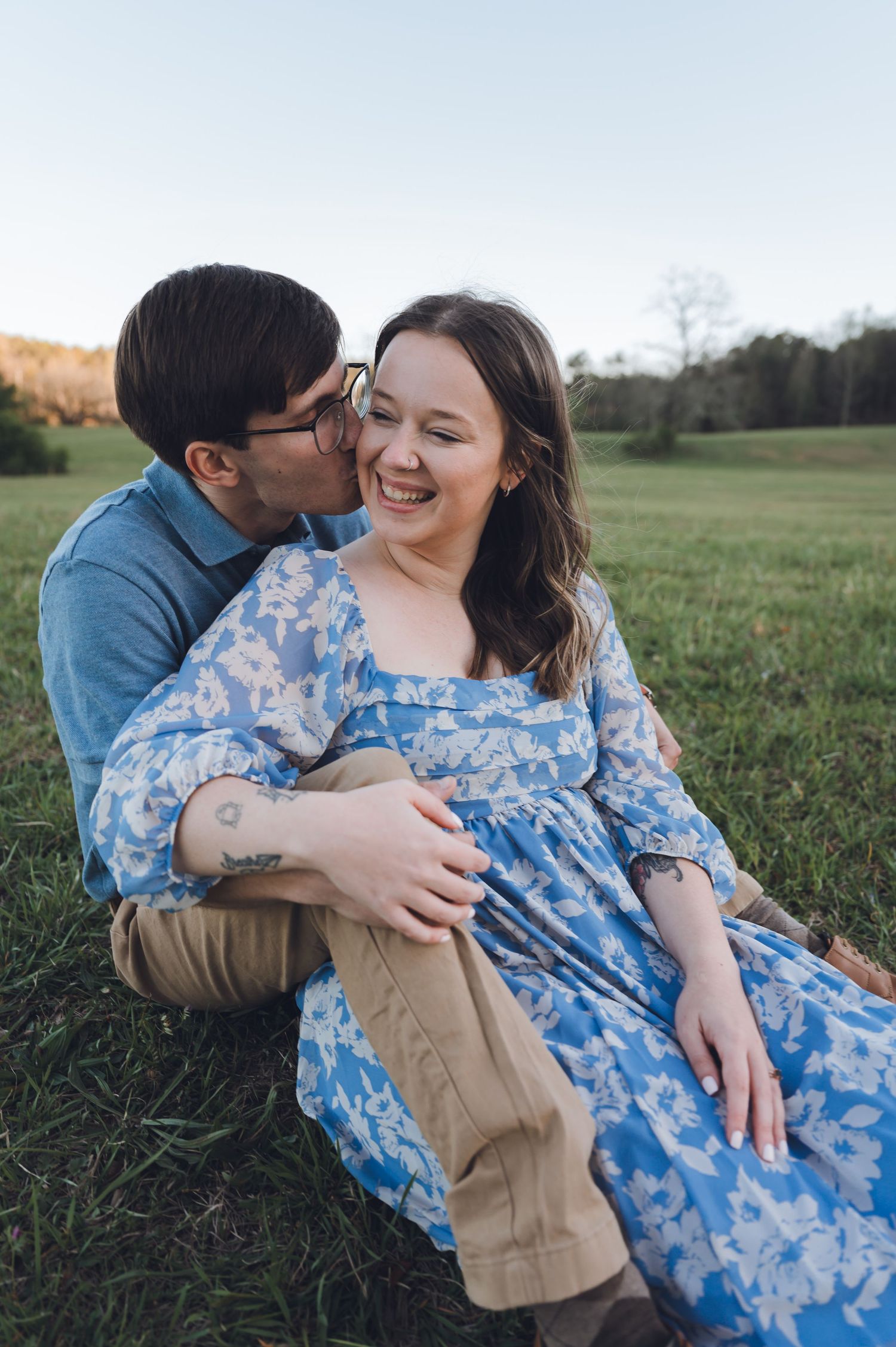 A couple sits in a green field, man kissing woman's cheek as she laughs, wearing a blue floral dress.