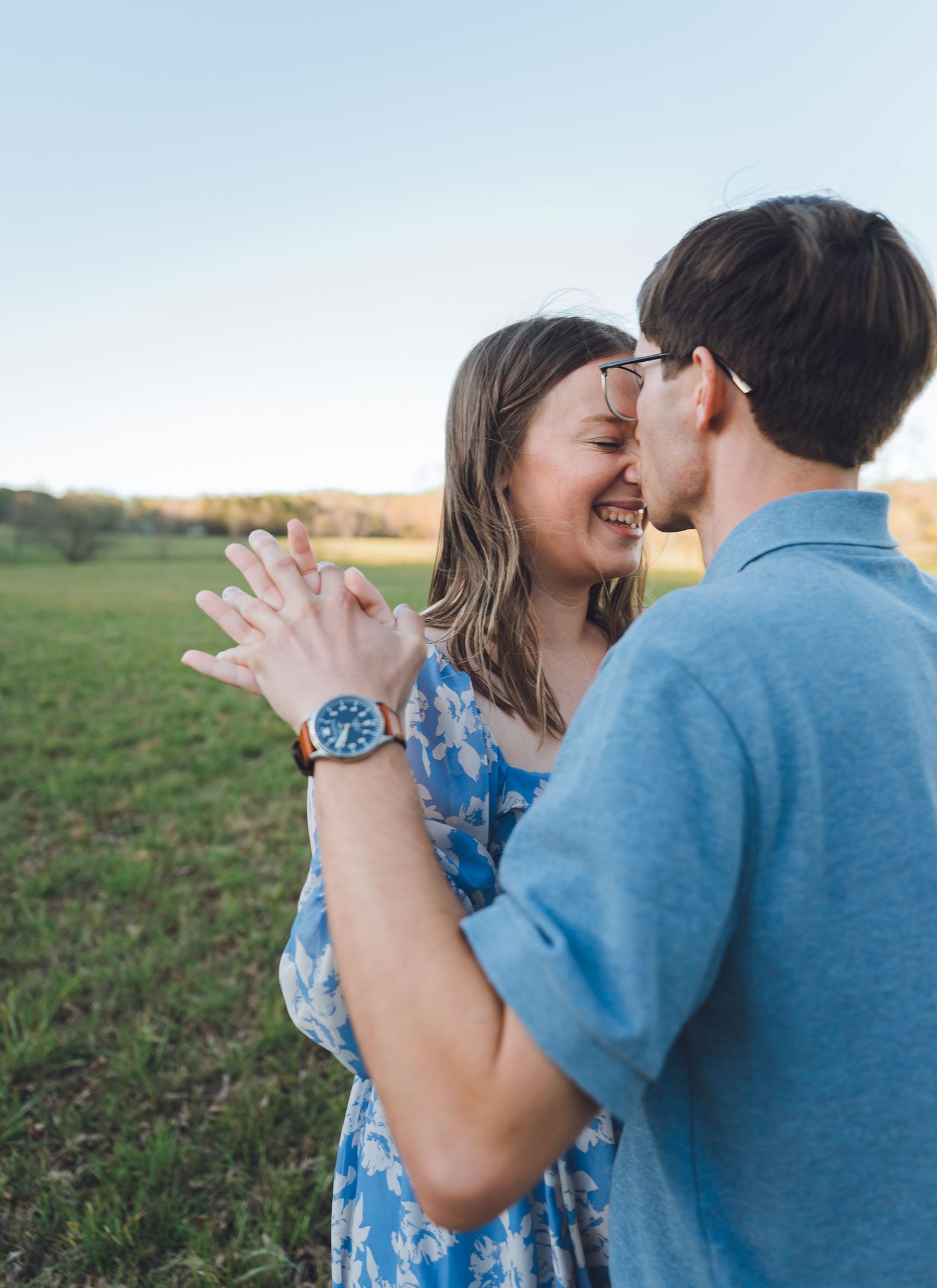A joyful couple embraces in a green field at golden hour, the woman laughing and showing her engagement ring.