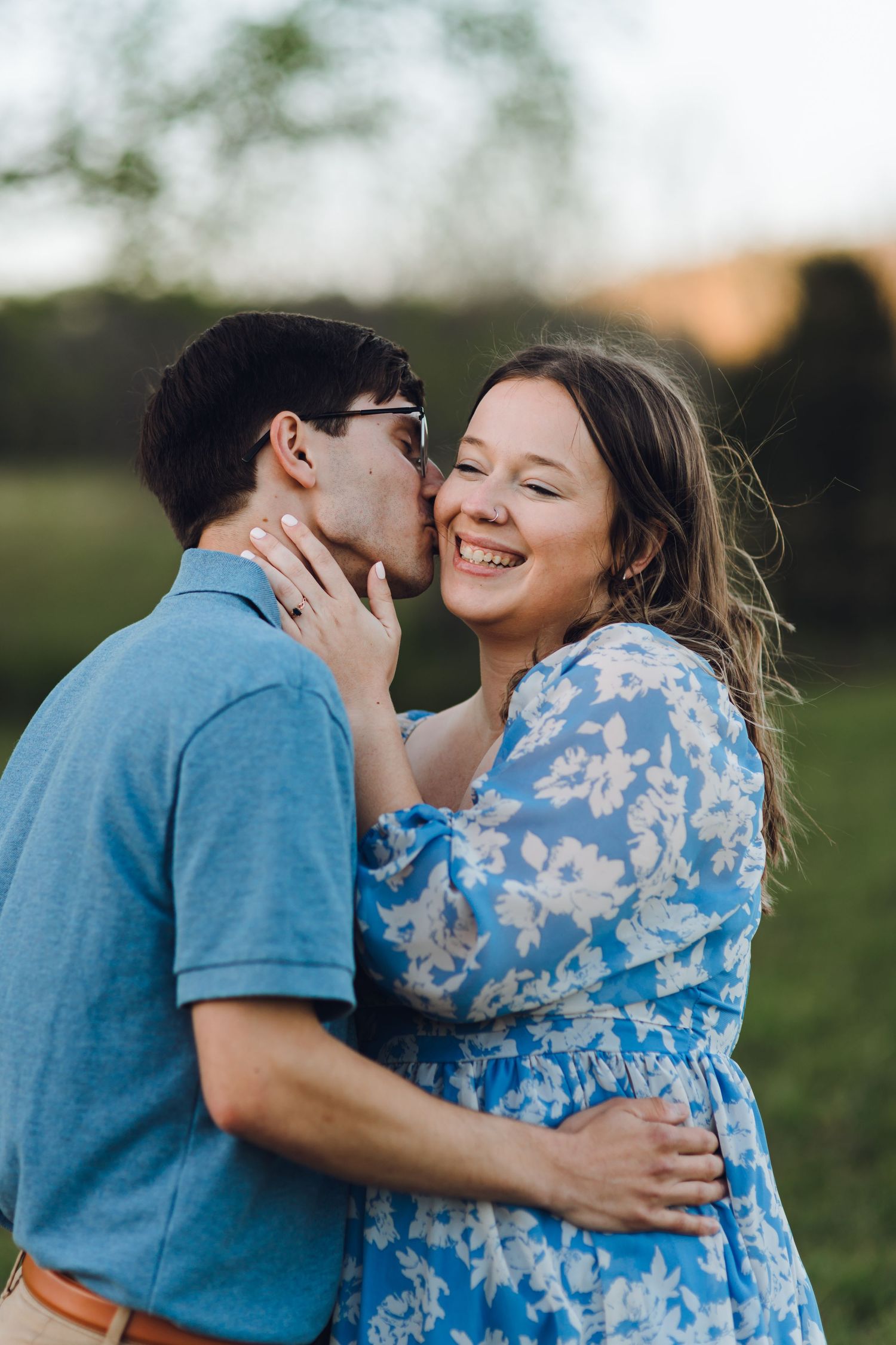 Couple embracing outdoors, man kissing woman's cheek as she smiles joyfully, both in blue outfits in a green field.