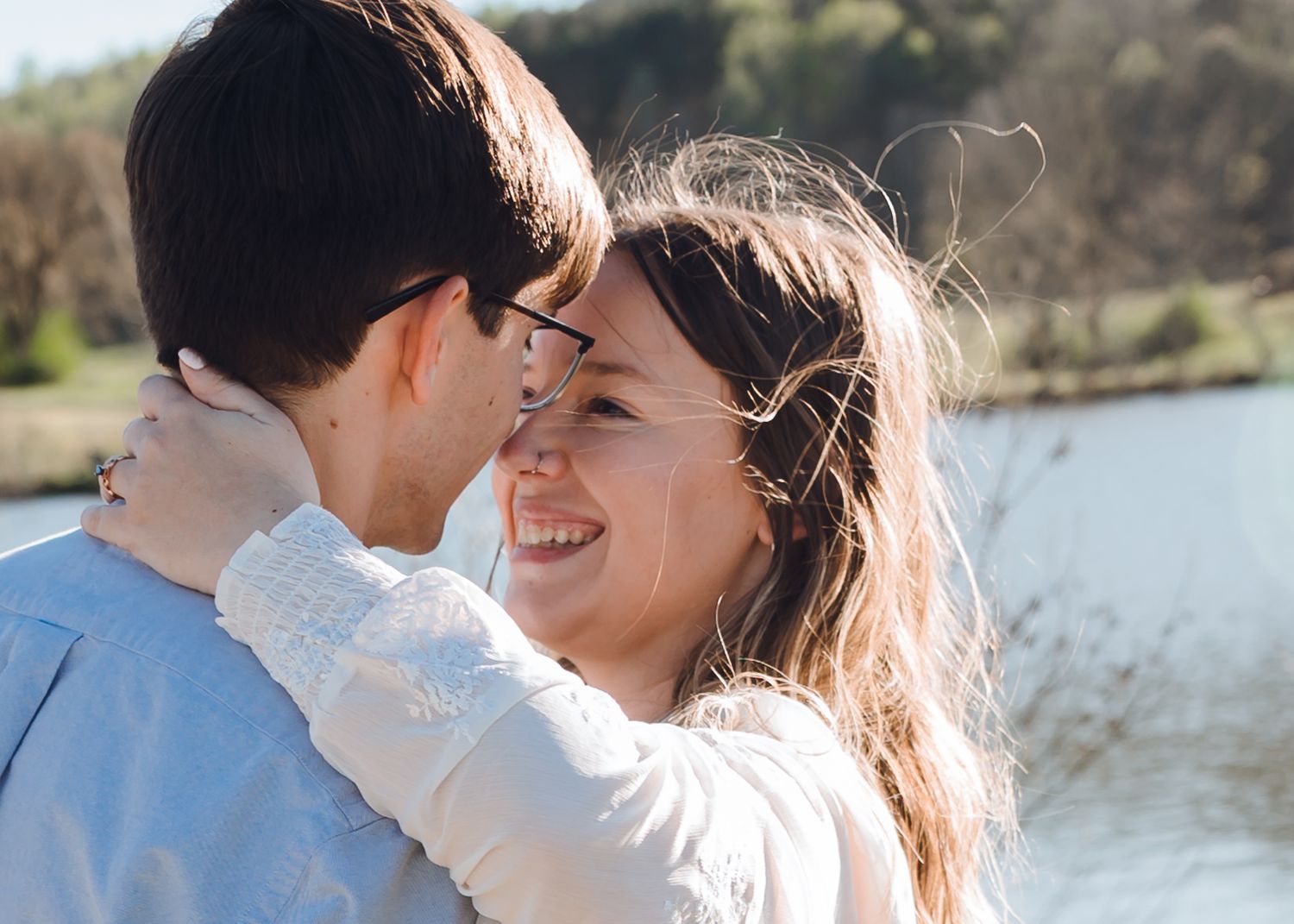 A smiling couple embraces outdoors near a lake on a sunny day, sharing a romantic moment.