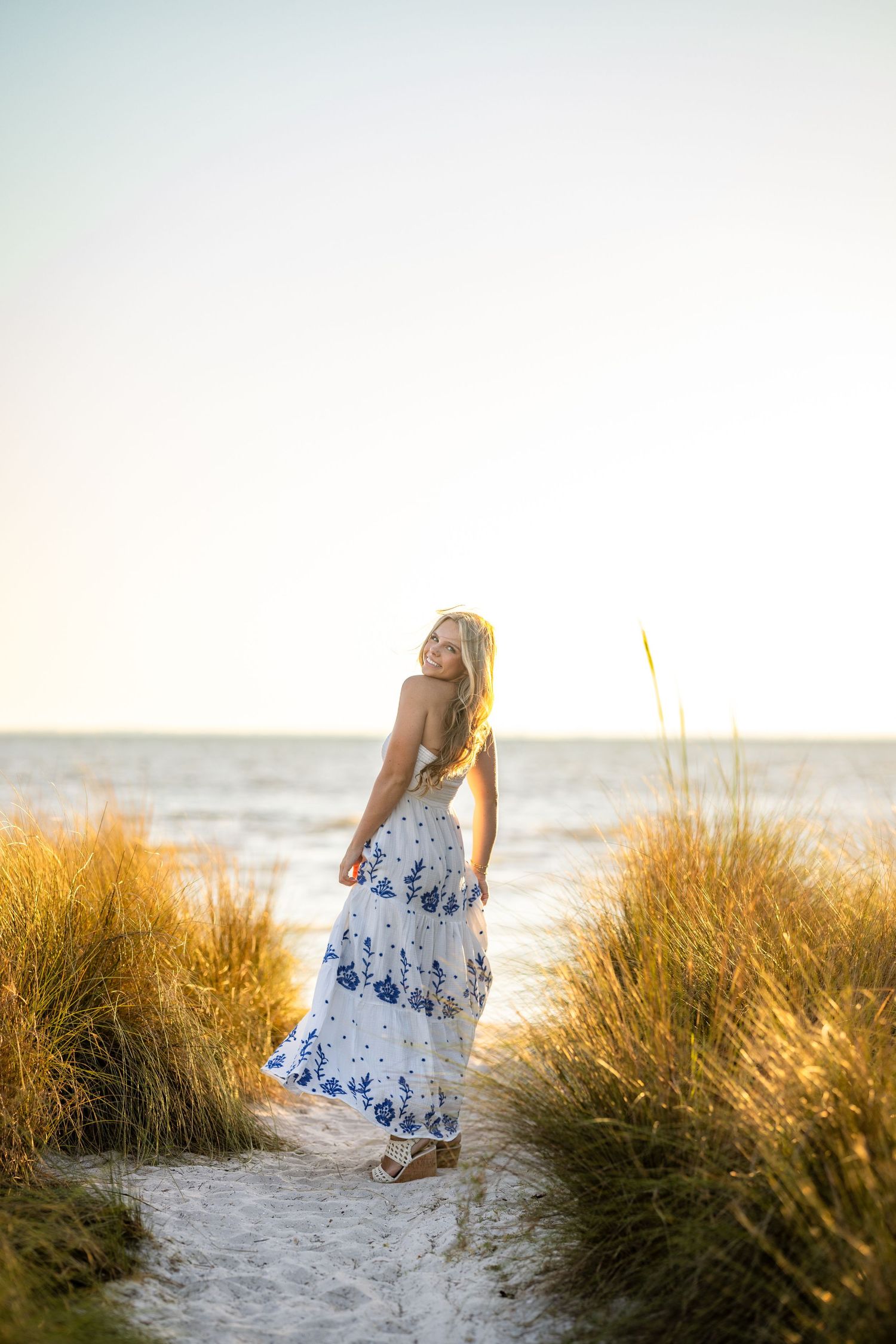 Young blonde woman in white floral dress smiling on wooden boardwalk surrounded by lush green trees.