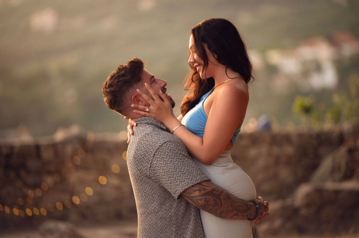 Couple sharing a tender moment outdoors, pregnant woman in white dress embraced by tattooed partner at golden hour.