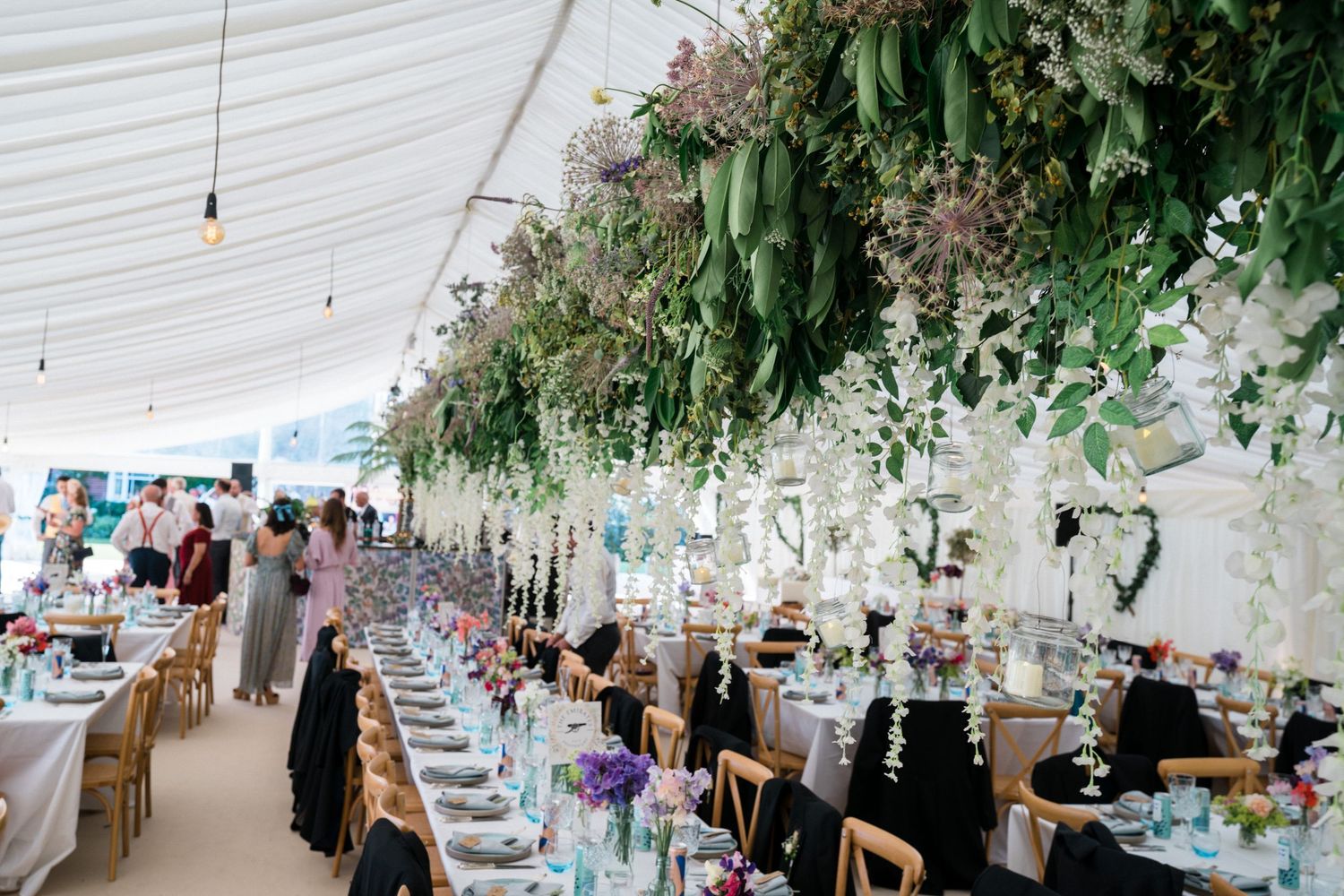 Elegant wedding reception marquee adorned with lush greenery ceiling installations and long banquet tables with floral centerpieces.