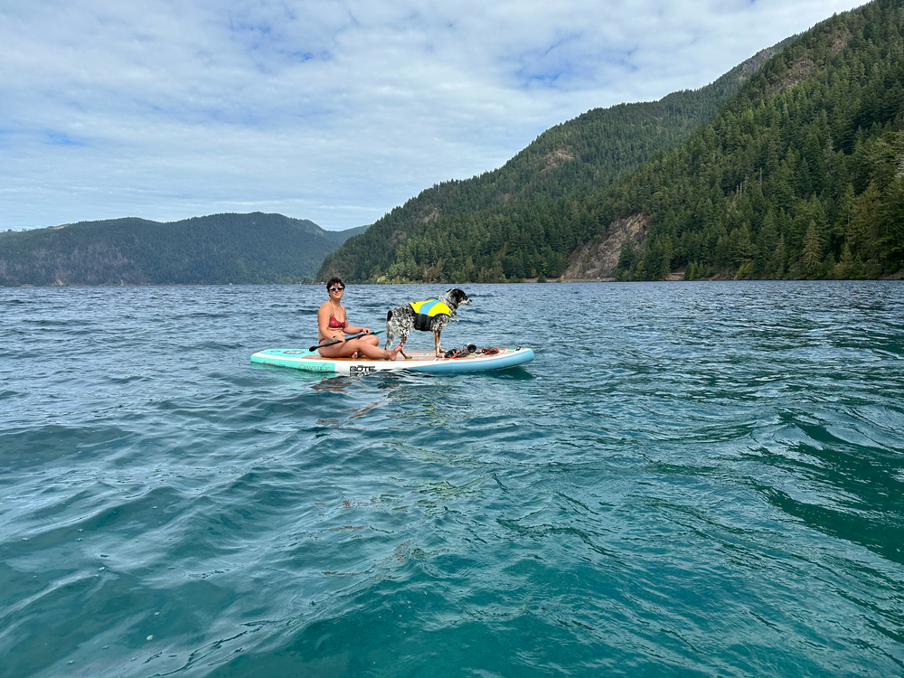 A woman paddle boards with her dog at an alpine lake.