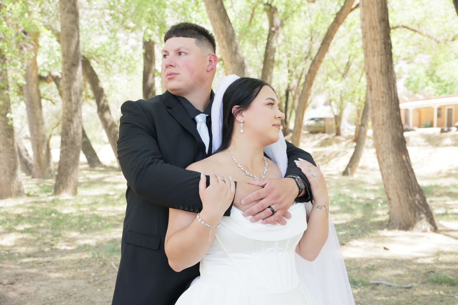 A groom in a black suit embraces his bride in a white dress outdoors among trees at a wedding ceremony.