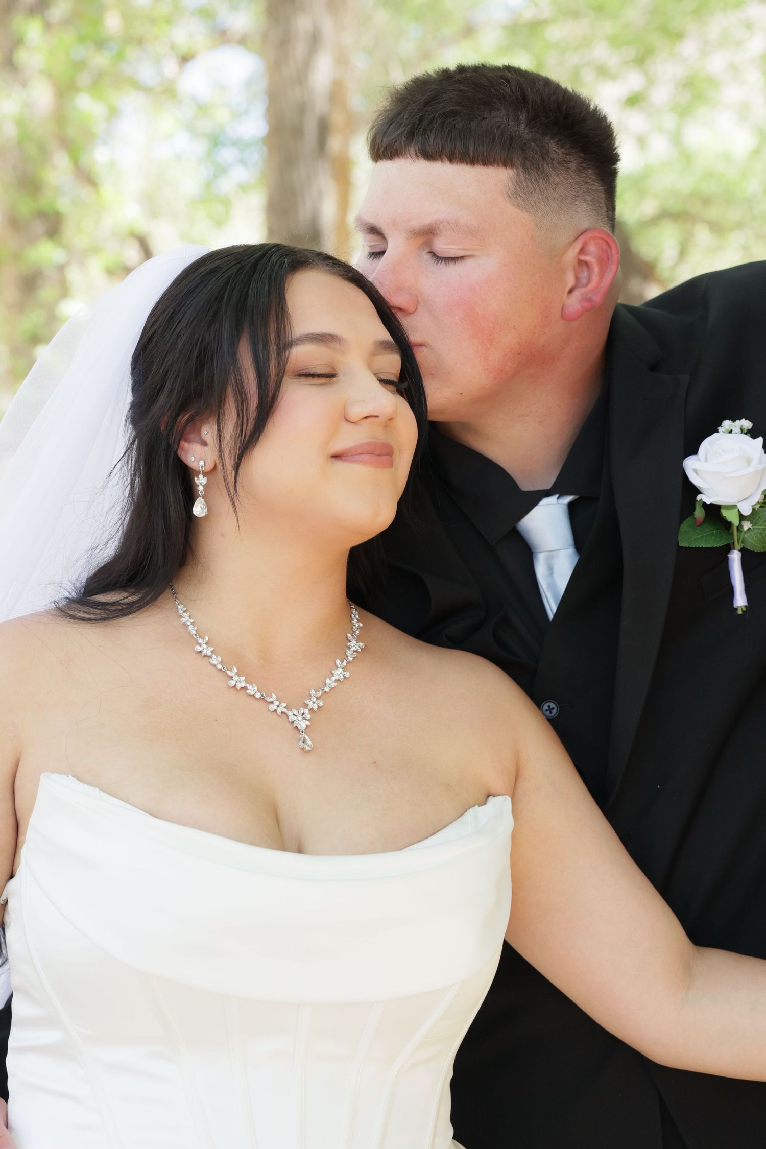 Groom in black tuxedo kissing bride on cheek, bride wearing white strapless gown and diamond necklace outdoors.