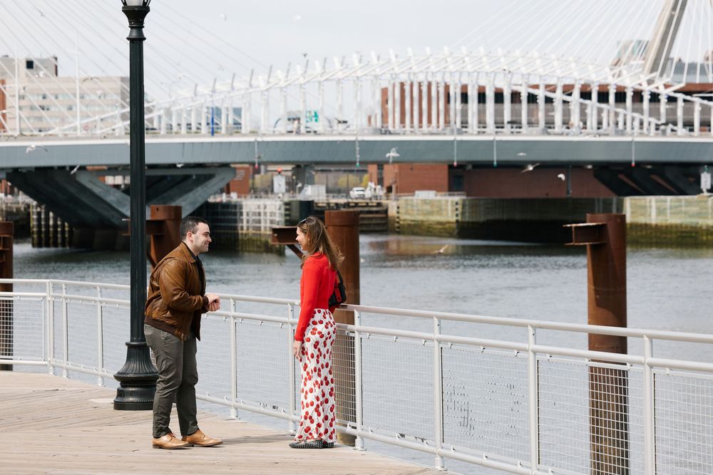 Couple standing on a waterfront boardwalk near a bridge, man in brown jacket and woman in red top with floral skirt.