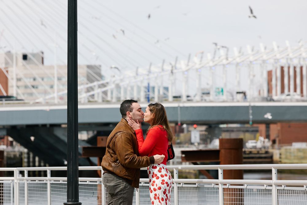 Couple sharing a romantic kiss on a waterfront bridge with a suspension bridge backdrop in an urban setting.