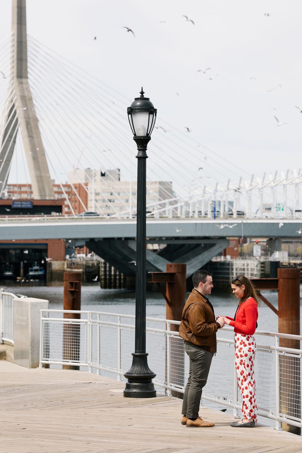 A couple stands by a lamppost on a waterfront dock with the Zakim Bridge in Boston visible behind them.