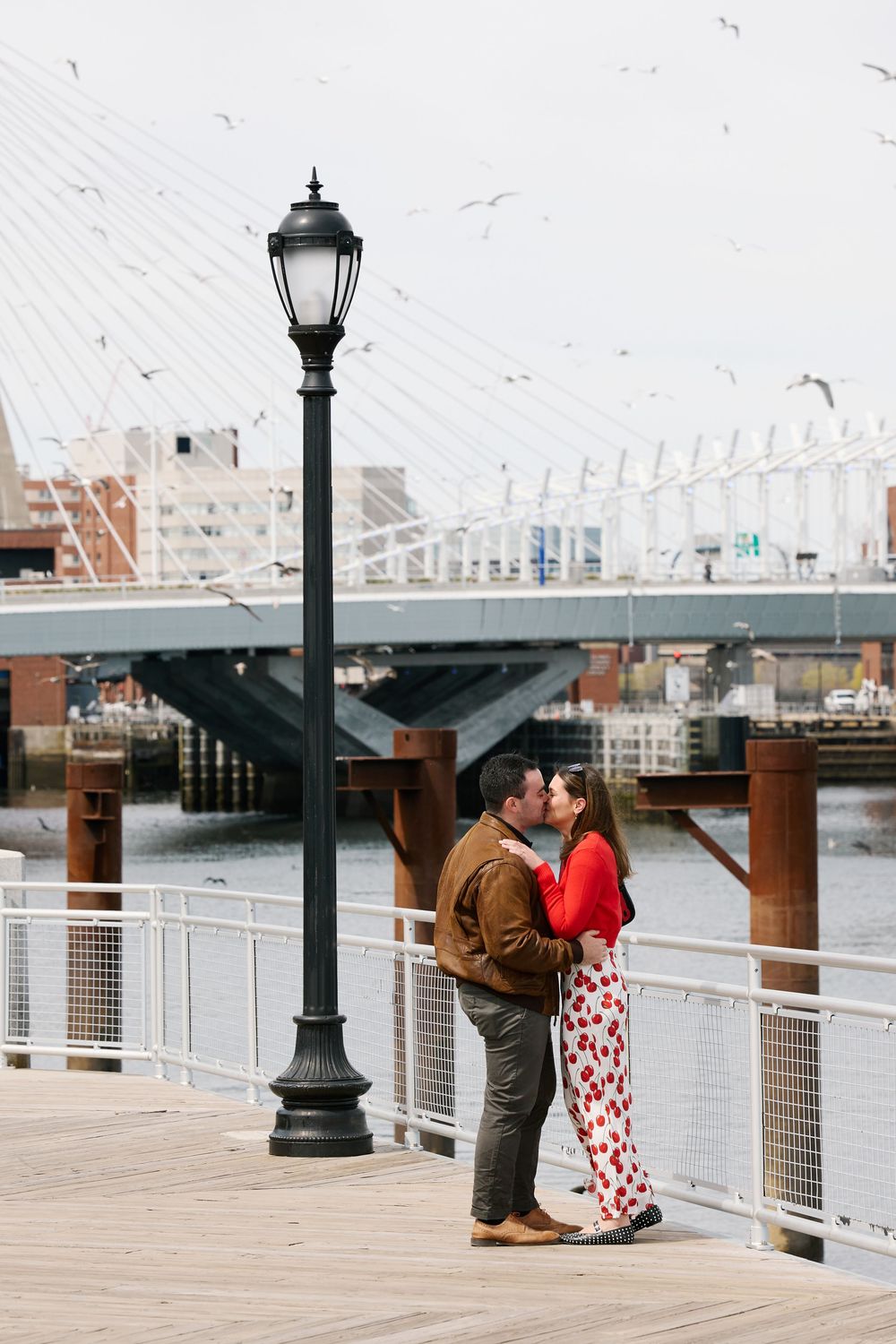 A couple shares a romantic kiss beside a black lamppost on a Boston waterfront dock with a cable-stayed bridge backdrop.