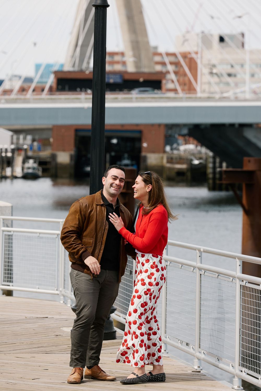 Couple smiling at each other on a waterfront boardwalk, woman in red top and floral skirt, man in brown leather jacket.