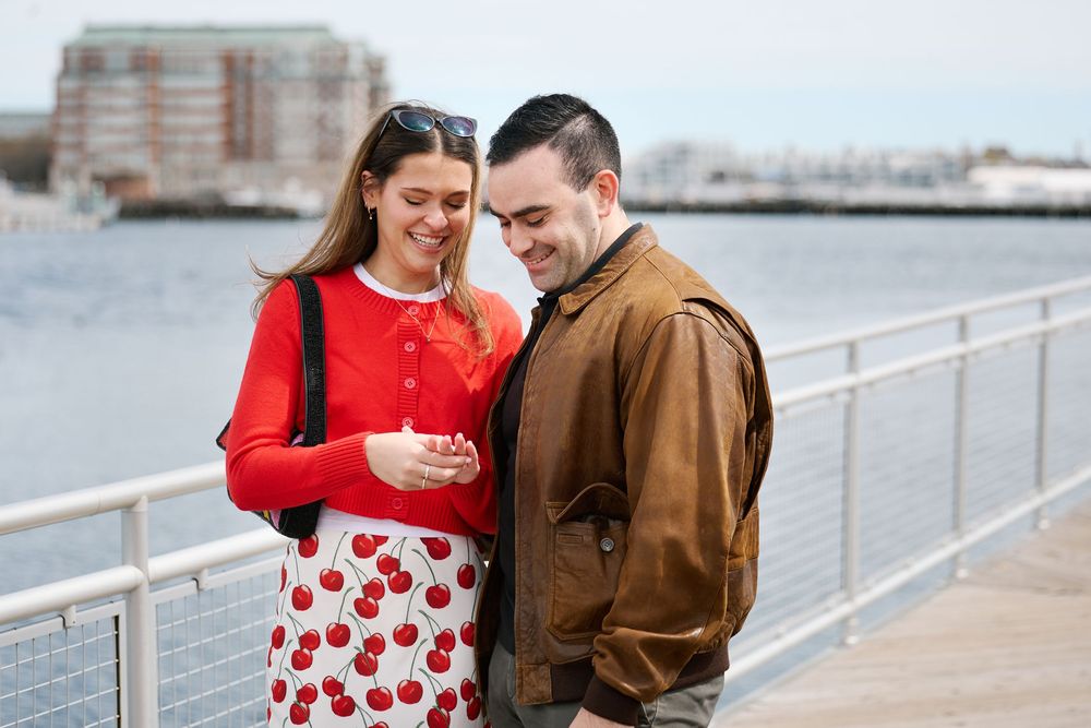 Couple smiling and looking at phone together on waterfront pier, woman in red cardigan and cherry print skirt.