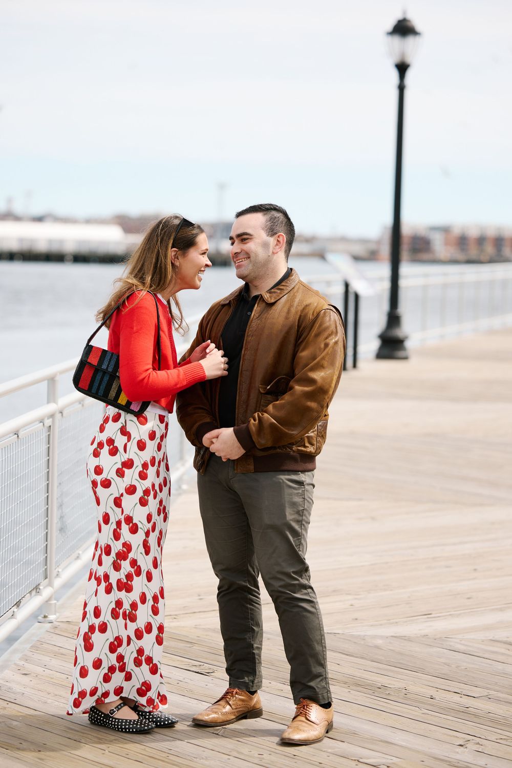 Happy couple embracing on a wooden boardwalk by the water, woman in floral maxi skirt and red cardigan.