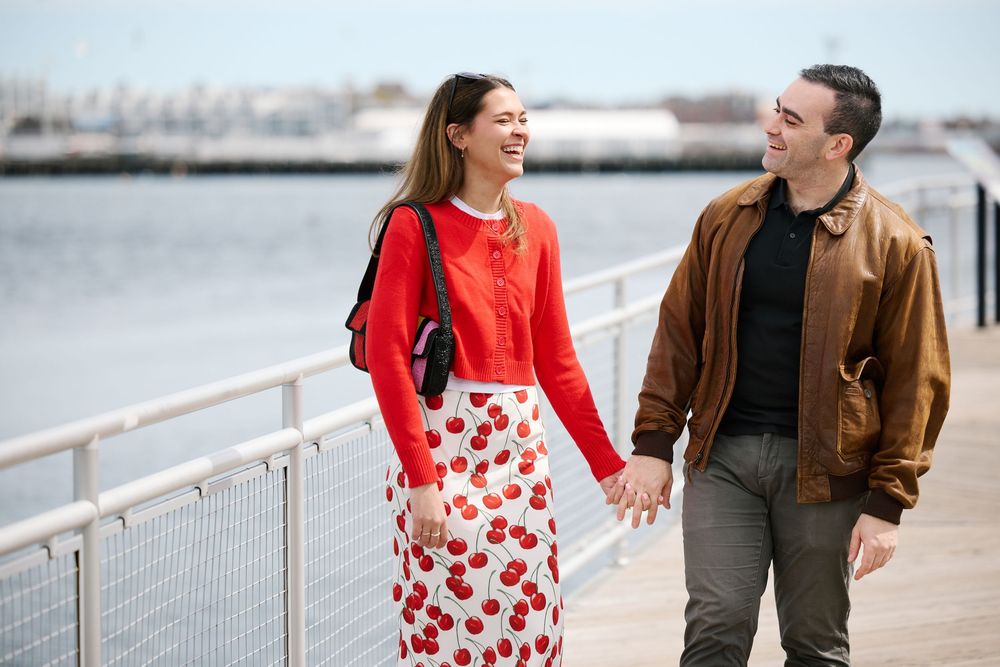 Happy couple holding hands and walking on a waterfront boardwalk, woman in red cardigan and floral skirt, man in brown jacket.