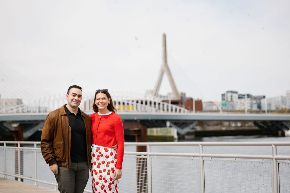 Couple smiling together on Boston waterfront with Zakim Bridge in background on a cloudy day.