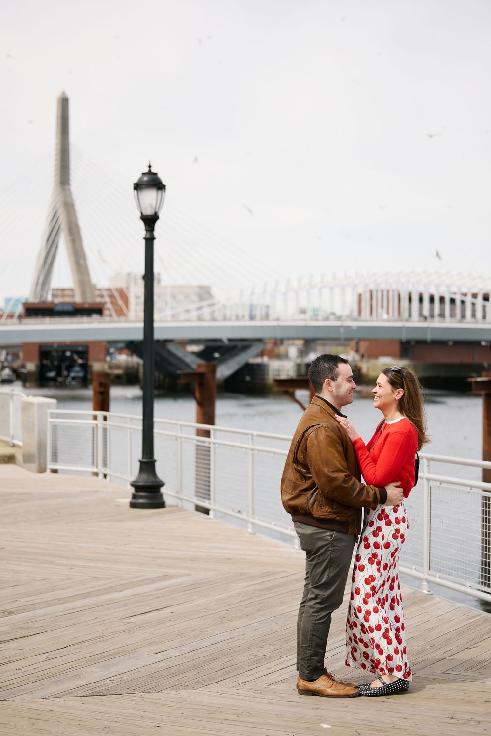 Romantic couple holding each other on a harbor pier with the Zakim Bridge visible in the background.