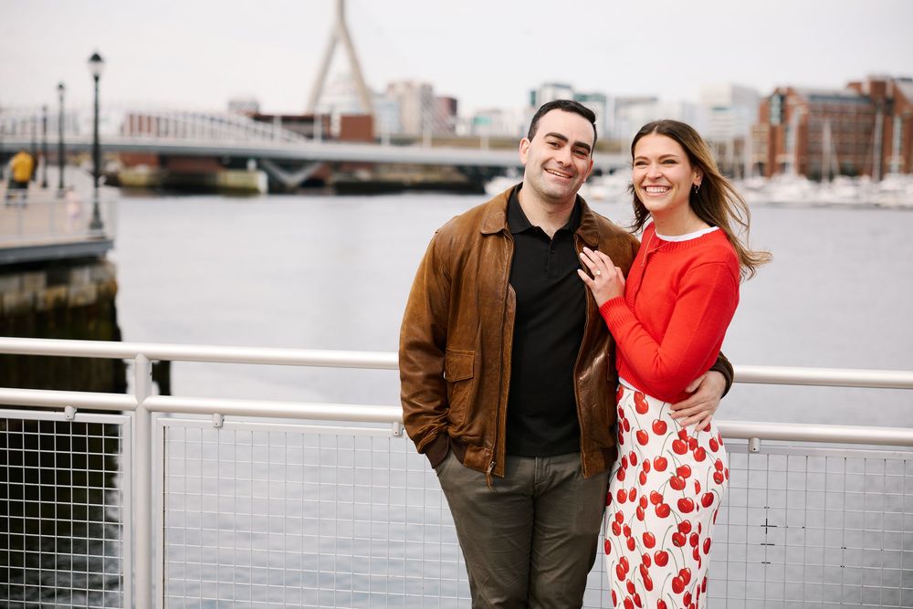 Couple smiling together on a waterfront bridge in Boston, woman in red top, man in brown jacket.