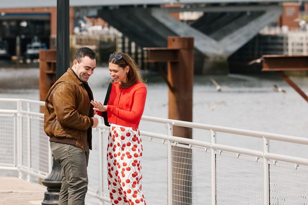 Couple smiling and looking at phone together on waterfront promenade, woman in red floral dress, man in brown jacket.
