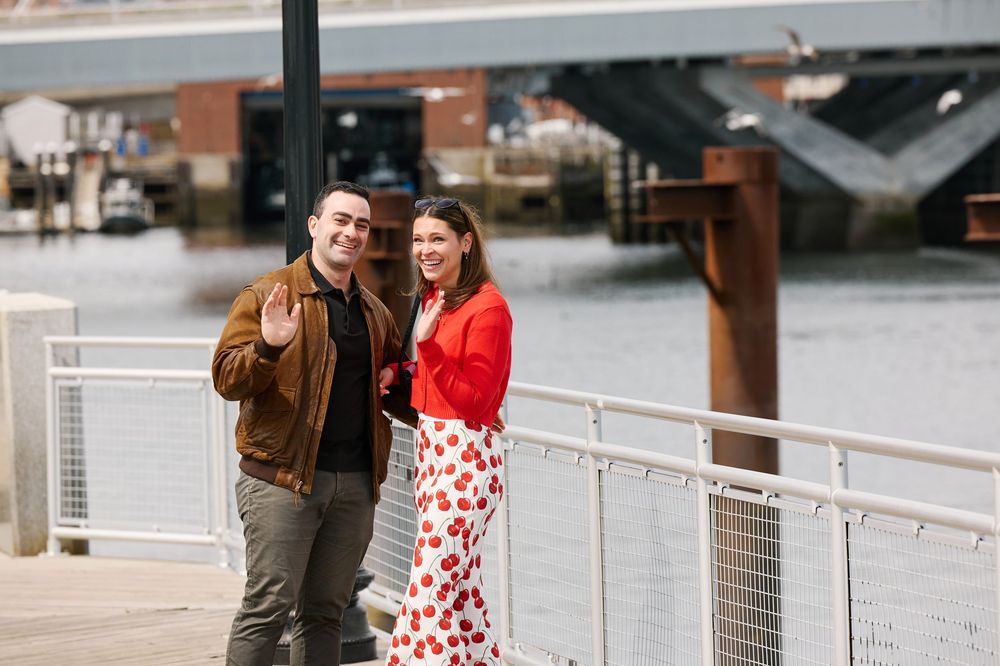Couple smiling on a waterfront dock, woman in floral skirt and red top, man in brown jacket giving thumbs up.